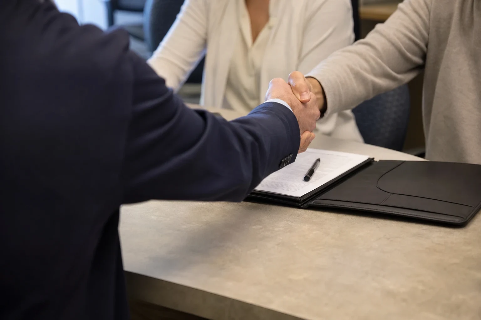 Over-the-shoulder view of an agent in a navy blazer shaking hands with a client across a desk, an open black folder with papers and a pen between them