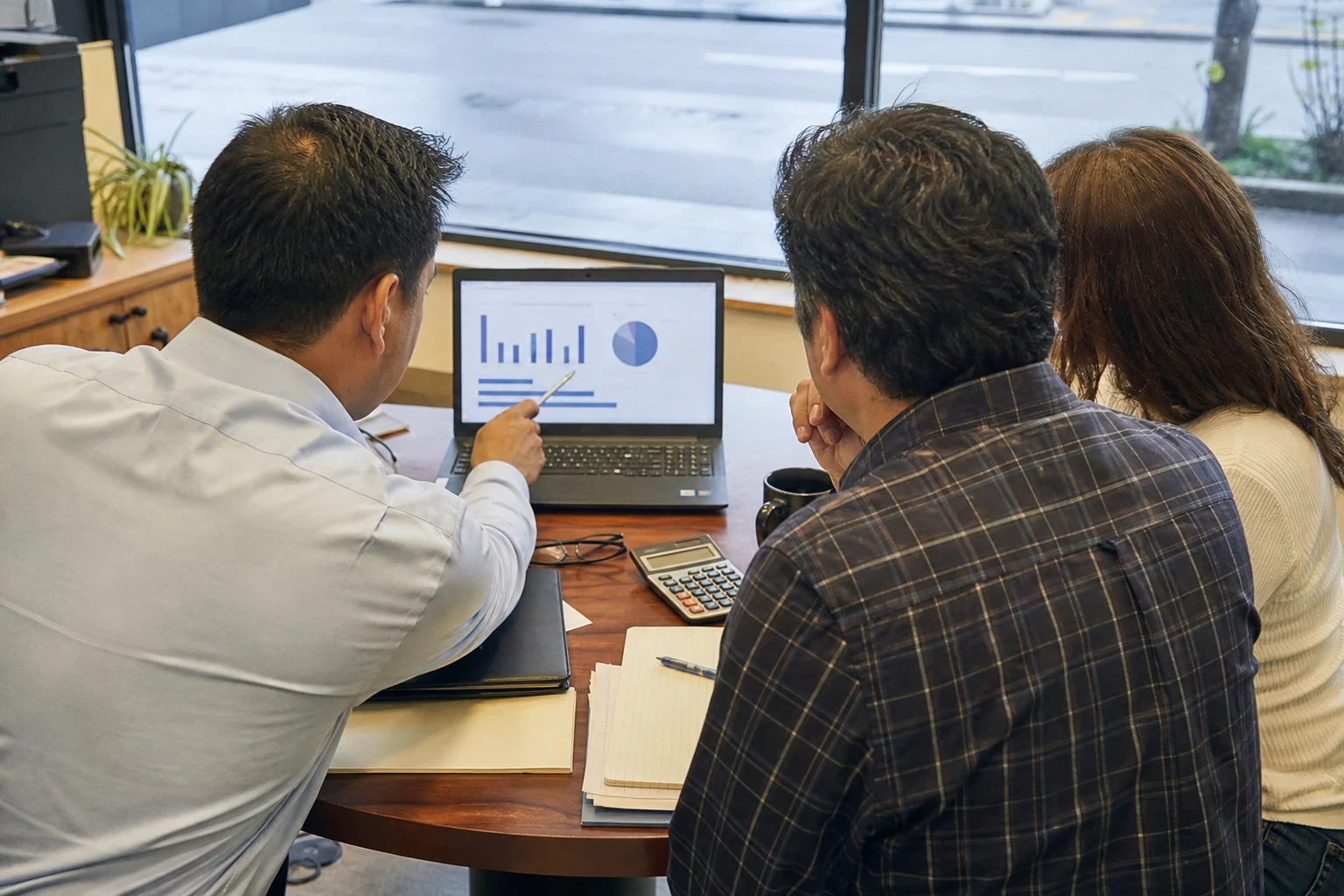 Agent in a light blue shirt points at charts on a laptop while a man in a plaid shirt and a woman look on at a round table by a storefront window.